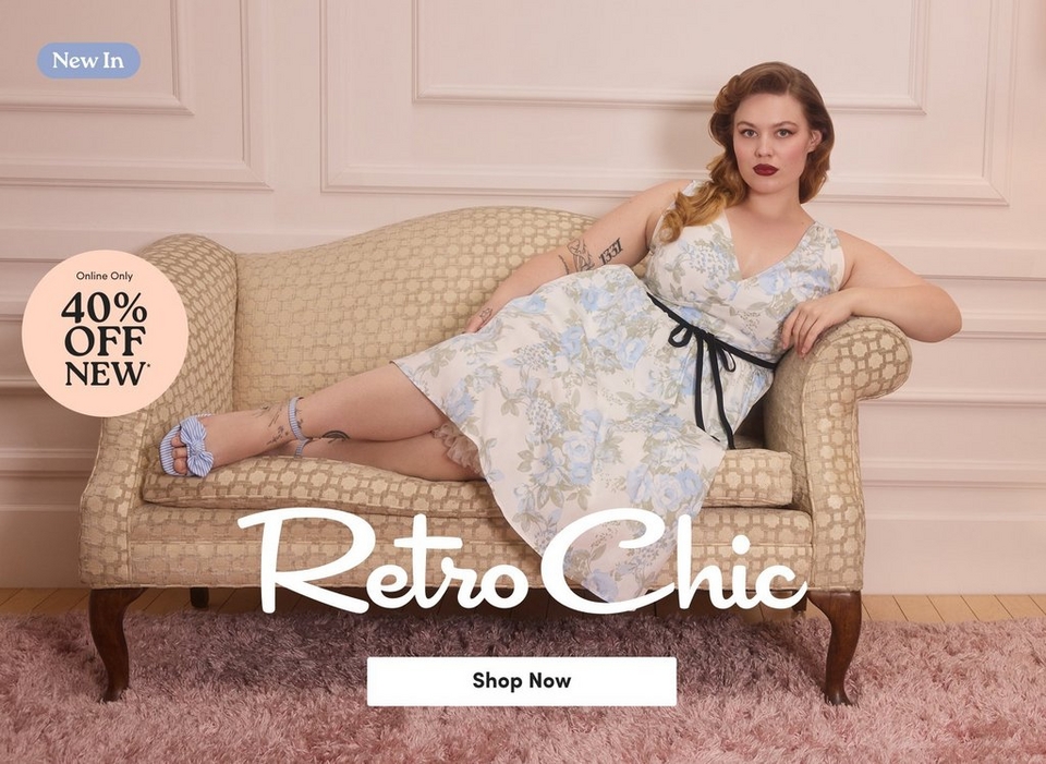 A promotional banner for the 'Retro Chic' collection featuring a 'New In' label in a blue pill-shaped badge in the upper left corner and a peach circular badge reading 'Online Only 40% Off New' on the left side. A woman with auburn hair styled in a vintage wave and side braid, wearing dark red lipstick and tattoos on her arms, reclines on a tan geometric-patterned loveseat. She wears a sleeveless V-neck midi dress in a cream, blue, and sage green floral print with a black ribbon tie at the waist, paired with light blue striped sandals with bow detail. The loveseat sits on a pink fluffy rug against a blush pink paneled wall with light wood flooring visible at the edges. The text 'Retro Chic' appears in large white script across the lower portion of the image above a white 'Shop Now' button.