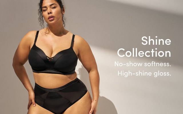A promotional banner for the 'Shine Collection' with the tagline 'No-show softness. High-shine gloss.' in white text on the right side. A woman with dark curly hair stands against a warm gray background with soft directional light and shadow, leaning slightly forward with one hand at her side. She wears a black underwire bra with satin and mesh paneling and a small decorative cutout detail at the center front, paired with matching black high-waist briefs with geometric mesh and satin paneling.