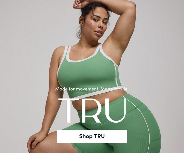 'A promotional banner for 'TRU' with the tagline 'Made for movement. Made for life.' and a white 'Shop TRU' button overlaid on a photo. A woman with dark hair pulled up and small hoop earrings sits against a light gray background with one arm raised overhead. She wears a sage green ribbed sports bra crop top with white trim detail and matching sage green ribbed biker shorts with white piping.'