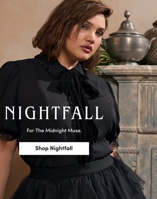 A promotional banner for 'Nightfall' with the tagline 'For The Midnight Muse.' and a white 'Shop Nightfall' button overlaid on a photo. A woman with short dark brown wavy hair, a nose ring, and defined eye makeup looks to the side while seated near a stone mantelpiece with ornate silver vessels and greenery in a warm beige-toned setting. She wears a black short-sleeve blouse with puff sleeves, ruffled bib front, velvet bow tie at the neck, and lace cuff detail, paired with a black tiered tulle skirt.