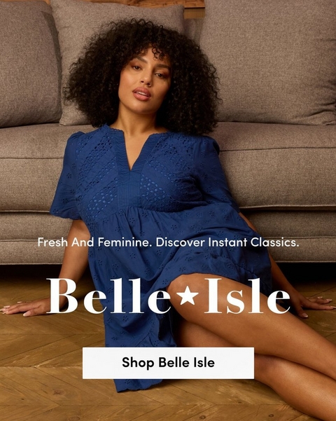 A promotional banner for 'Belle Isle' with the tagline 'Fresh And Feminine. Discover Instant Classics.' and a white 'Shop Belle Isle' button overlaid on a photo. A woman with voluminous natural curly dark hair sits on a herringbone wood floor leaning against a gray upholstered sofa. She wears a cobalt blue short-sleeve V-neck midi dress with intricate allover broderie anglaise eyelet detailing and a tiered skirt.'