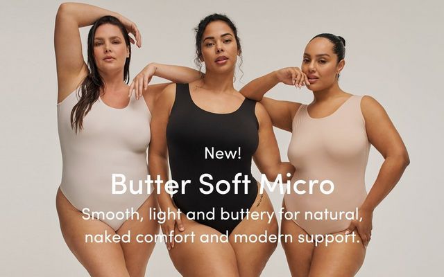 A promotional banner for 'New! Butter Soft Micro' with the tagline 'Smooth, light and buttery for natural, naked comfort and modern support.' in white text overlaid across a photo of three women standing close together against a light gray background. The woman on the left has long dark hair with one arm raised overhead and wears a white smooth seamless scoop-neck thong bodysuit. The woman in the center has dark curly hair pulled up and one hand raised near her chin, wearing a black smooth seamless scoop-neck thong bodysuit. The woman on the right has dark hair in a bun and one hand raised near her chin, wearing a blush pink smooth seamless scoop-neck thong bodysuit.