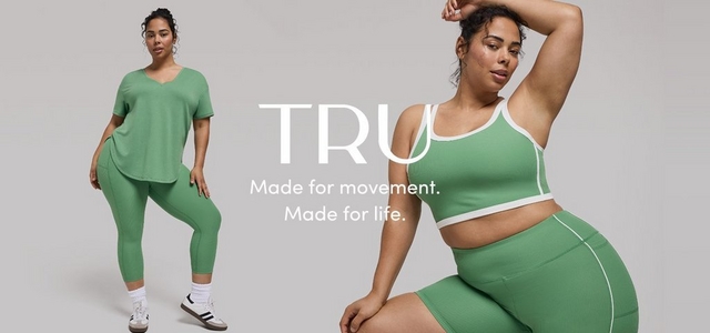 A promotional banner for 'TRU' with the tagline 'Made for movement. Made for life.' in white text overlaid across two side-by-side photos against a light gray background. On the left, a woman with dark hair in a bun, small hoop earrings, and white sneakers with a dark sole stands wearing a sage green V-neck oversized short-sleeve top with matching sage green cropped leggings and white ankle socks. On the right, the same woman is shown in a closer crop with one arm raised overhead, wearing a sage green ribbed sports bra crop top with white trim and matching sage green ribbed biker shorts with white piping.