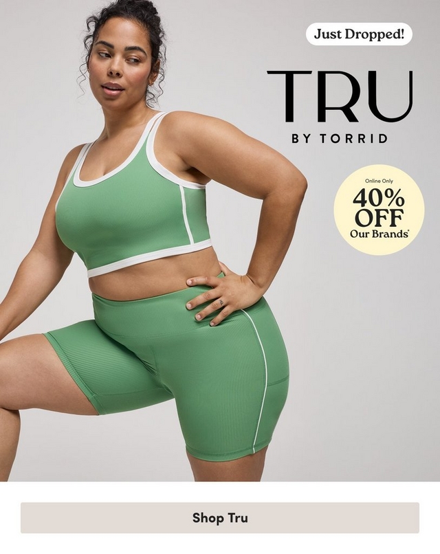 'Promotional banner for Tru by Torrid on a light gray background. A plus-size woman with medium-deep skin tone and dark curly hair pulled up poses with one knee raised and her hand on her hip, looking to the side. She is wearing a green ribbed sports bra crop top with white trim and double straps, paired with matching green ribbed biker shorts with a white piping detail. She wears small gold hoop earrings and a ring. In the upper right, a white rounded badge reads 'Just Dropped!' above large bold dark text reading 'TRU' with smaller text below reading 'BY TORRID'. Below that, a pale yellow circle badge with dark text reads 'Online Only 40% OFF Our Brands' with an asterisk. At the bottom is a light gray rectangular button with bold dark text reading 'Shop Tru'.'