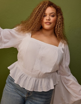 'A plus-size woman with medium skin tone and long voluminous curly auburn and honey-highlighted hair poses against a flat olive green painted wall. She is wearing a white long-sleeve square-neck cropped blouse with lace trim detailing down the front panels, a ruffled peplum hem with lace trim, and wide flared sleeves with lace cuff accents, paired with light wash blue jeans. She is looking directly at the camera with one arm slightly extended.'