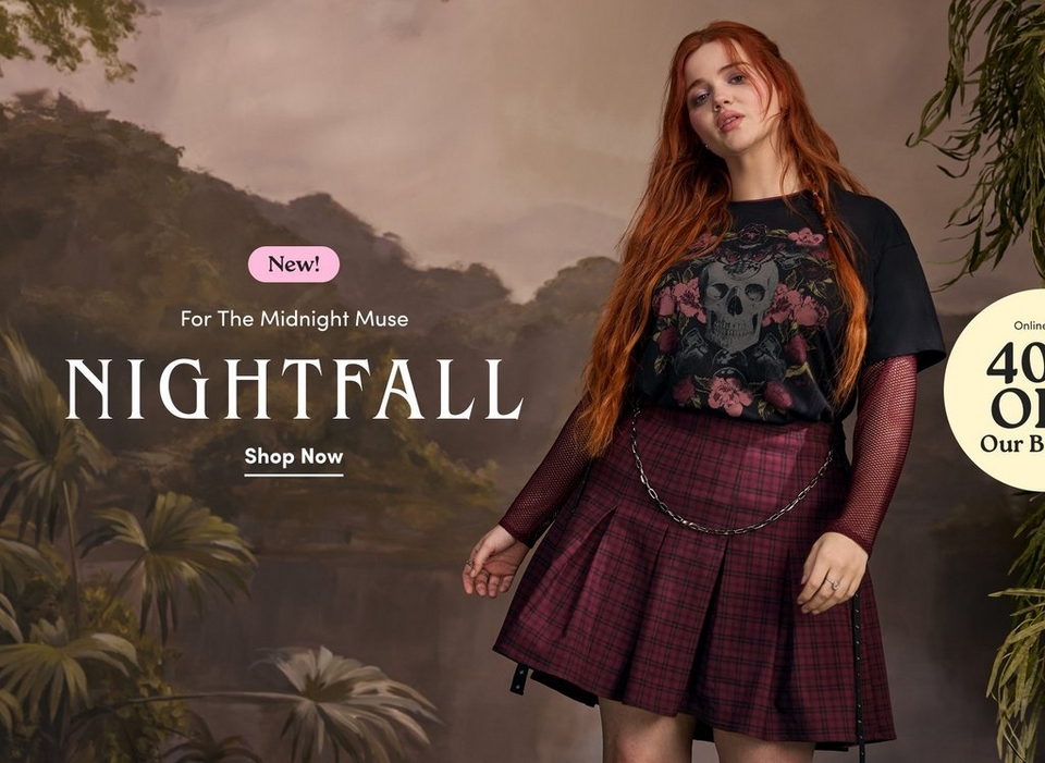 'Promotional banner for Torrid Nightfall collection against a dark, moody painted backdrop depicting a misty jungle landscape with trees, tropical foliage, and a cloudy sky in muted brown and green tones. Hanging green vines are visible on the right edge. A plus-size woman with long wavy red hair and light skin stands in the center-right of the image, looking toward the camera with a subtle expression. She is wearing a black graphic t-shirt featuring a skull and floral rose design, layered over a long-sleeve black fishnet top, paired with a dark burgundy and black plaid pleated mini skirt with a silver chain belt and black studded strap detail. She wears rings on both hands. On the left side, a small pink rounded badge reads 'New!' above smaller white text reading 'For The Midnight Muse', followed by large white serif text reading 'NIGHTFALL'. Below that, bold white underlined text reads 'Shop Now'. On the far right, a partially visible pale yellow circle badge shows dark text reading '40% OFF Our B' with the rest cut off.'