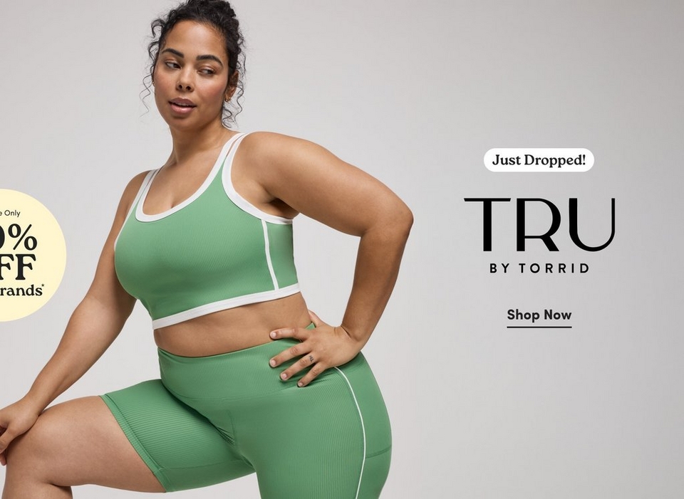 'Promotional banner for Tru by Torrid on a light gray background. A plus-size woman with medium-deep skin tone and dark curly hair pulled up poses with one knee raised and her hand on her hip, looking to the side. She is wearing a green ribbed sports bra crop top with white trim and double straps, paired with matching green ribbed biker shorts with a white piping detail. She wears small gold hoop earrings and a ring. On the left edge, a partially visible pale yellow circle badge shows dark text reading 'e Only 0% FF rands' with an asterisk, indicating the full badge reads 'Online Only 40% OFF Our Brands'. On the right side, a white rounded badge reads 'Just Dropped!' above large bold dark text reading 'TRU' with smaller text below reading 'BY TORRID'. Below that, bold dark underlined text reads 'Shop Now'.'