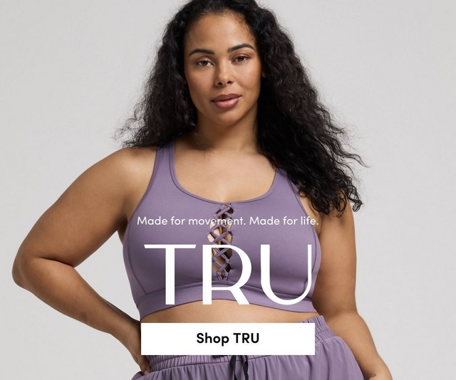 'Promotional banner for Tru by Torrid against a solid light gray background. A plus-size woman with medium skin tone and long dark curly hair looks directly at the camera with a neutral expression, one hand on her hip. She is wearing a lavender purple sports bra with a criss-cross lace-up detail at the center front, and matching lavender jogger shorts with a black drawstring waist. Smaller white text reads 'Made for movement. Made for life.' above large bold white text reading 'TRU'. At the bottom is a white rectangular button with bold dark text reading 'Shop TRU'.'