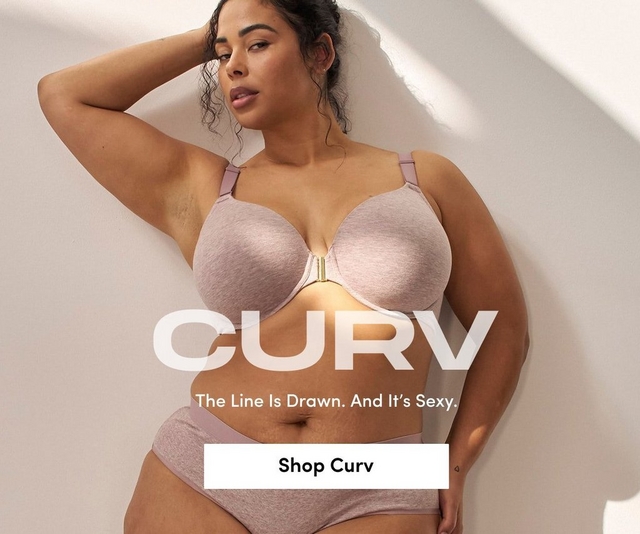 'Promotional banner for Torrid CURV against a warm beige background with soft light and shadow. A plus-size woman with medium skin tone and dark hair pulled up in a loose bun leans against a wall with one hand raised behind her head, looking to the side. She is wearing a dusty mauve heathered t-shirt bra with a gold front clasp detail and matching high-waist bikini briefs, with a small heart tattoo visible on her hip. Large bold white text overlaid on the lower portion reads 'CURV' with smaller white text above reading 'The Line Is Drawn. And It's Sexy.' At the bottom is a white rectangular button with bold dark text reading 'Shop Curv'.'
