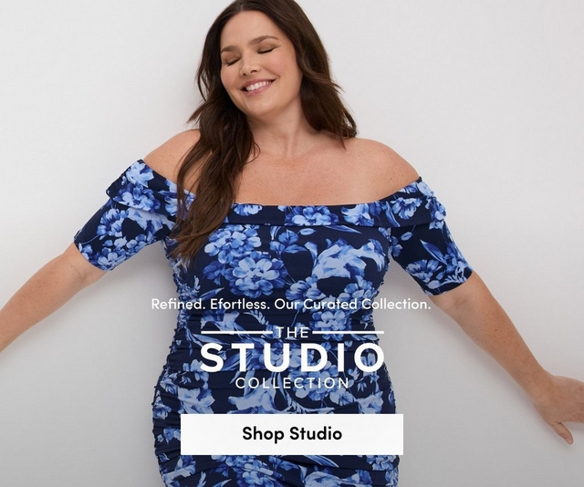 'Promotional banner for Torrid featuring The Studio Collection against a clean white background. A plus-size woman with light skin tone and long dark brown hair poses with eyes closed and arms extended outward, smiling softly. She is wearing a navy blue and periwinkle floral off-the-shoulder ruched midi dress with short puff sleeves and a fitted ruched skirt. Smaller white text reads 'Refined. Effortless. Our Curated Collection.' above bold white text flanked by horizontal lines reading 'THE STUDIO COLLECTION'. At the bottom is a white rectangular button with bold dark text reading 'Shop Studio'.'