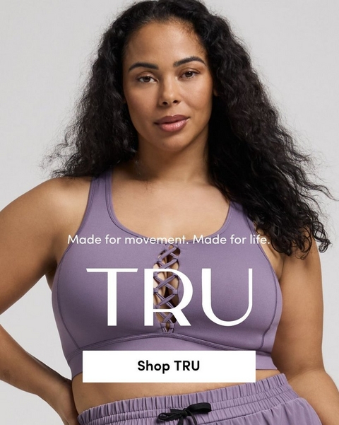 'Promotional banner for Tru by Torrid against a solid light gray background. A plus-size woman with medium skin tone and long dark curly hair looks directly at the camera with a neutral expression, one hand on her hip. She is wearing a lavender purple sports bra with a criss-cross lace-up detail at the center front, and matching lavender jogger shorts with a black drawstring waist. Smaller white text reads 'Made for movement. Made for life.' above large bold white text reading 'TRU'. At the bottom is a white rectangular button with bold dark text reading 'Shop TRU'.'