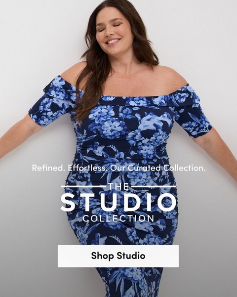 'Promotional banner for Torrid featuring The Studio Collection against a clean white background. A plus-size woman with light skin tone and long dark brown hair poses with eyes closed and arms extended outward, smiling softly. She is wearing a navy blue and periwinkle floral off-the-shoulder ruched midi dress with short puff sleeves and a fitted ruched skirt. Smaller white text reads 'Refined. Effortless. Our Curated Collection.' above bold white text flanked by horizontal lines reading 'THE STUDIO COLLECTION'. At the bottom is a white rectangular button with bold dark text reading 'Shop Studio'.'