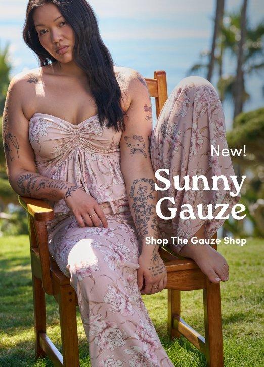 'Promotional banner for Torrid featuring Sunny Gauze. A plus-size woman with medium skin tone and long straight dark hair sits on a wooden chair outdoors on a grassy area with palm trees and blue sky in the background. She is wearing a pink and cream floral gauze strapless top with a ruched and knotted center front detail, paired with matching floral gauze wide-leg pants. Extensive tattoos are visible on both arms and legs. In the upper right, small dark text reads 'New!' above larger dark serif text reading 'Sunny Gauze' with small dark underlined text below reading 'Shop The Gauze Shop'.'
