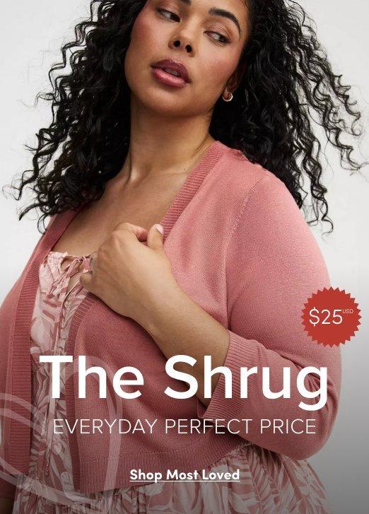 A promotional banner with a light gray background. A woman with long dark curly hair and a small stud earring is shown from the chest up, looking slightly downward. She wears a soft pink knit shrug cardigan open at the front over a floral printed top. One hand rests on her chest. A red circular badge in the upper right reads '$25' with a superscript asterisk. Overlaid white text in the lower portion reads 'The Shrug' in large letters, with 'EVERYDAY PERFECT PRICE' in smaller text below it. At the bottom is a white rectangular button with dark text reading 'Shop Most Loved'.