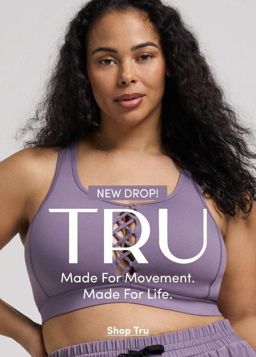 'Promotional banner for Tru by Torrid against a light gray background. A plus-size woman with medium skin tone and long dark curly hair looks directly at the camera with a neutral expression. She is wearing a lavender purple sports bra with a criss-cross lace-up detail at the center front and matching lavender jogger shorts with a drawstring waist. A small white rounded badge reads 'NEW DROP!' above large bold white text reading 'TRU' with smaller white text below reading 'Made For Movement. Made For Life.' At the bottom, small dark underlined text reads 'Shop Tru'.'