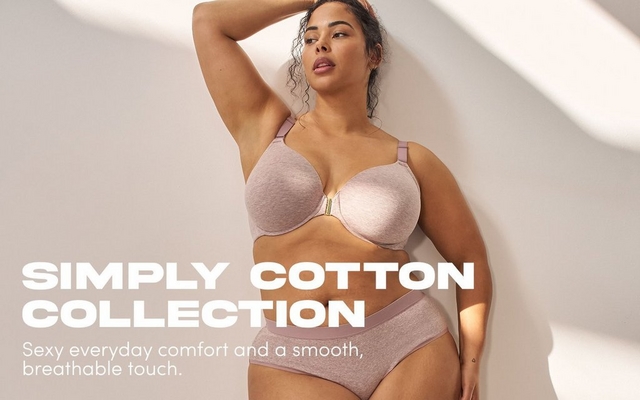 'A promotional banner for the Simply Cotton Collection featuring a woman with dark curly hair pulled loosely up, posing with one arm raised and bent behind her head against a warm off-white wall with soft diagonal shadow. She wears a heathered blush pink cotton bra with smooth molded cups and a small gold clasp at the center front, paired with matching blush pink high-waist cotton briefs with a dusty mauve waistband. Bold white text in the lower left reads 'SIMPLY COTTON COLLECTION' with smaller text below reading 'Sexy everyday comfort and a smooth, breathable touch.''