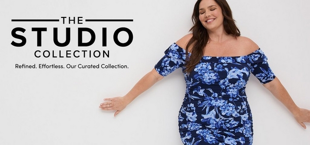 'Promotional banner for Torrid featuring The Studio Collection against a clean white background. A plus-size woman with light skin tone and long dark brown hair poses with eyes closed and arms extended outward, smiling softly. She is wearing a navy blue and periwinkle floral off-the-shoulder ruched midi dress with short puff sleeves and a fitted ruched skirt. On the left side, bold dark text flanked by horizontal lines reads 'THE STUDIO COLLECTION' with smaller dark text below reading 'Refined. Effortless. Our Curated Collection.'.'