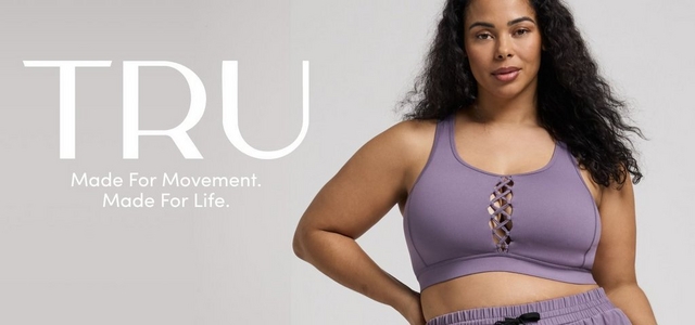 'Promotional banner for Tru by Torrid against a solid light gray background. A plus-size woman with medium skin tone and long dark curly hair stands with one hand on her hip, looking directly at the camera. She is wearing a lavender purple sports bra with a criss-cross lace-up detail at the center front, and dark gray jogger shorts with a drawstring waist. On the left side, large bold white text reads 'TRU' with smaller white text below reading 'Made For Movement. Made For Life.'.'