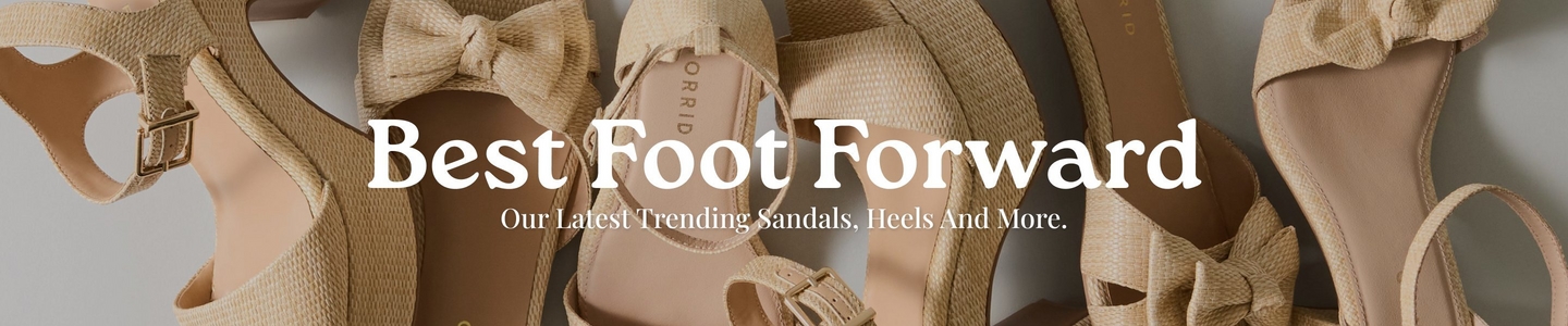 'Promotional banner for Torrid featuring a close-up flat lay of multiple neutral-toned raffia and woven textured women's sandals and heels arranged overlapping each other against a light gray background. The shoes include styles with bow details, ankle straps with gold buckles, block heels, and platform soles, all in warm beige and tan tones. The Torrid brand name is visible on the insole of one shoe. Overlaid in large bold white serif text is 'Best Foot Forward' with smaller white text below reading 'Our Latest Trending Sandals, Heels And More.'.'