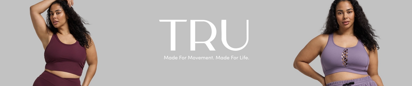 'Promotional banner for Tru by Torrid against a solid light gray background. Two plus-size women with medium skin tone and long dark curly hair are positioned on either side of the banner. On the left, a woman poses with one arm raised above her head, wearing a deep burgundy ribbed sports bra crop top and matching high-waist shorts. On the right, a woman stands with one hand on her hip, wearing a lavender purple sports bra with a criss-cross lace-up detail at the center front, and dark gray jogger shorts with a drawstring waist. In the center, large bold white text reads 'TRU' with smaller white text below reading 'Made For Movement. Made For Life.'.'
