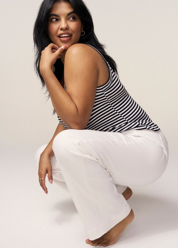 'A woman with long straight black hair crouches low against a light warm gray background, turning her head to look over her shoulder with a playful smile. She wears small gold hoop earrings, a navy and white horizontal stripe sleeveless tank top with a racerback silhouette, and white wide-leg pants. Her hand rests near her chin with fingers lightly curled, and her other hand rests on her knee. She wears a gold ring on one finger and is barefoot.'