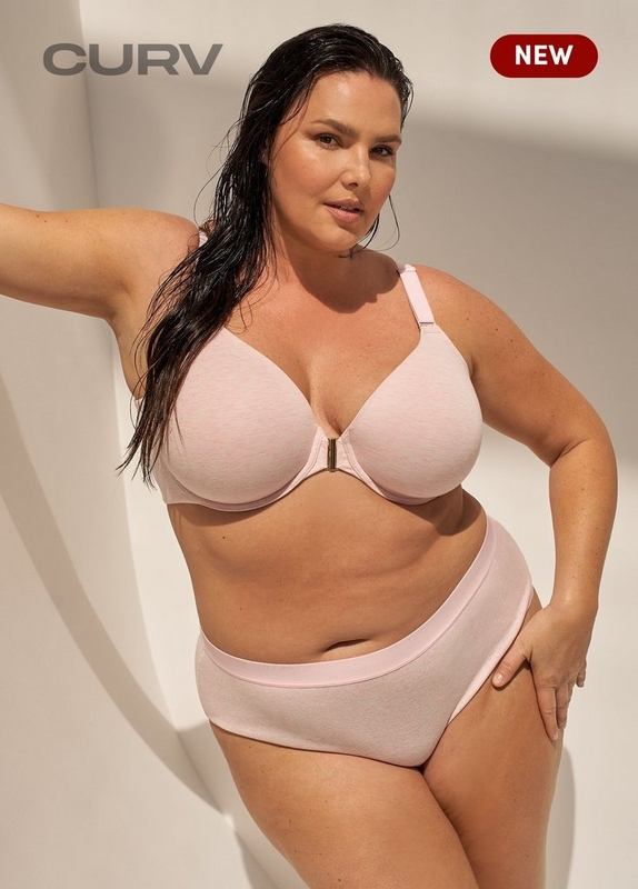 'A plus-size woman with long dark wet hair poses leaning against a warm beige wall with soft diagonal light and shadow. She wears a matching blush pink cotton lingerie set consisting of a smooth molded-cup bra with wide adjustable straps and a small gold center-front clasp, paired with high-waist briefs featuring a slightly darker pink waistband. Gray text in the upper left reads 'CURV' and a dark red rounded badge in the upper right reads 'NEW' in white text.'