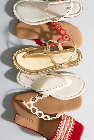 'A flat lay of five sandals arranged in an overlapping diagonal row on a light gray surface with soft shadows. From top to bottom: a cream leather t-strap thong sandal with gold hardware and the Torrid logo embossed on the insole; a red flat sandal with a chain-link strap detail featuring circular gold grommet accents and a tan footbed; a gold metallic t-strap thong sandal with a gold buckle closure and the Torrid logo embossed on the gold insole; a white flat thong sandal with a wide toe post strap and the Torrid logo embossed on the cream insole; and a white flat sandal with a chain-link strap featuring gold grommet accents, a tan footbed, and a partial view of a red espadrille slide with rope-wrapped platform detailing at the bottom edge.'