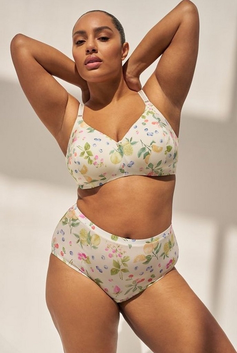 'A plus-size woman with her hair pulled back close to her head poses with both arms raised and hands clasped behind her head against a warm beige background with soft diagonal shadow. She wears a matching lingerie set in a cream base with a watercolor botanical print featuring lemons, blueberries, strawberries, pink flowers, and green leaves. The set consists of a wireless T-shirt style bra with a V-neckline and thin straps, and high-waist full-coverage briefs with a smooth waistband.'
