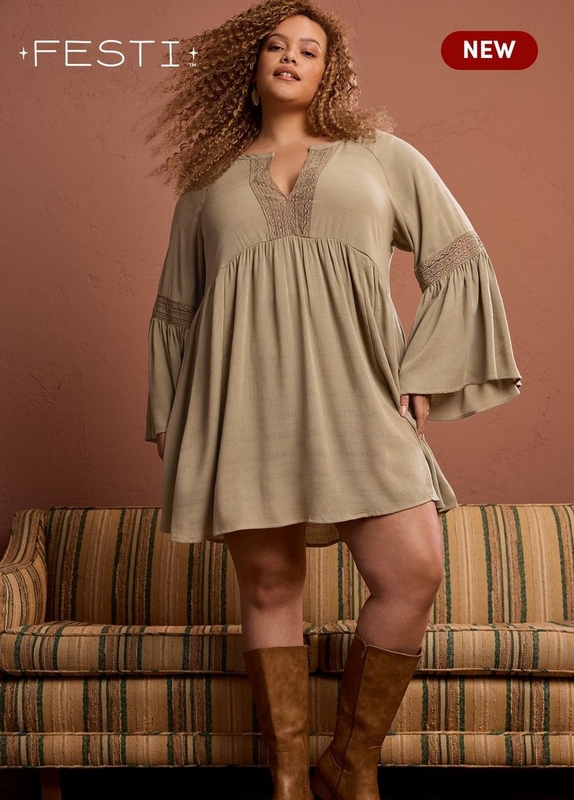 'A plus-size woman with long voluminous curly auburn hair poses standing on a striped green and tan upholstered sofa against a warm terracotta brown wall. She wears small gold earrings and a sage green babydoll mini dress with a V-neckline featuring lace trim detailing, an empire waist with gathered skirt, long bell sleeves with lace trim banding at the elbow, and a relaxed flowy silhouette. She is paired with tall tan leather knee-high boots. White text in the upper left reads '+FESTI+ TM' and a dark red rounded badge in the upper right reads 'NEW' in white text.'