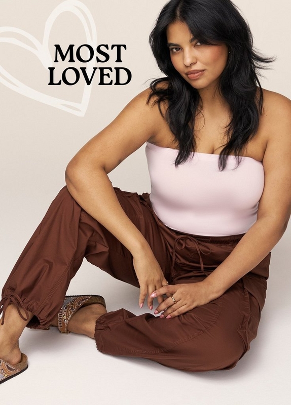 'A plus-size woman with long dark hair sits on the floor against a warm off-white background, leaning slightly to one side with her hands resting on her knees and gazing directly at the camera. She wears light pink nail polish and gold rings, a pale pink seamless strapless tube top, and rust brown wide-leg parachute pants with a drawstring waist, side cargo pockets, and tie-cuffed ankles. She wears tan flat sandals with beaded ankle strap detailing. A large outlined heart shape is visible in the upper left background. Bold dark text in the upper left reads 'MOST LOVED'.'