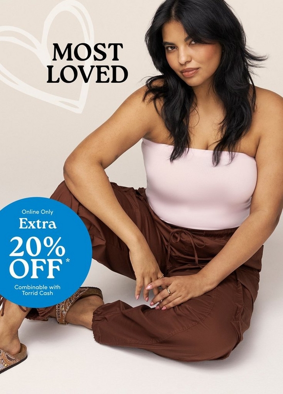 'A plus-size woman with long dark hair sits on the floor against a warm off-white background, leaning slightly to one side with her hands resting on her knees and gazing directly at the camera. She wears light pink nail polish and gold rings, a pale pink seamless strapless tube top, and rust brown wide-leg parachute pants with a drawstring waist and elastic cuffed ankles. She wears tan flat sandals with beaded ankle strap detailing. A large outlined heart shape is visible in the upper left background. Bold dark text in the upper left reads 'MOST LOVED'. A bright blue circle badge in the lower left contains white text reading 'Online Only' above 'Extra' and large text reading '20% OFF*' with smaller text below reading 'Combinable with Torrid Cash'.'
