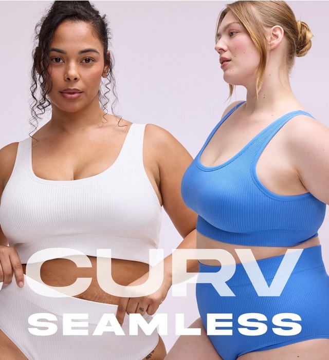 'A mobile promotional banner for Curv Seamless featuring two plus-size women against a soft lavender-gray background. On the left, a woman with dark curly hair pulled back looks directly at the camera wearing a white ribbed seamless scoop-neck sports bra crop top with matching high-waist seamless bikini briefs, and a small finger tattoo. On the right, a woman with strawberry blonde hair in a loose bun looks to the side wearing a periwinkle blue ribbed seamless scoop-neck racerback sports bra crop top with matching high-waist seamless shorts. Large semi-transparent text in the lower center reads 'CURV' above bold white text reading 'SEAMLESS'.'