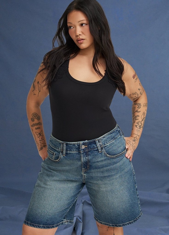 ''A plus-size woman with long dark hair and extensive tattoos covering both arms poses with both hands on her hips against a dark blue fabric backdrop. She wears a black ribbed scoop-neck sleeveless bodysuit with a small floral lace applique detail at the left shoulder, paired with medium-wash denim longline shorts with frayed hems, a zip fly, and five-pocket styling with gold hardware.'
