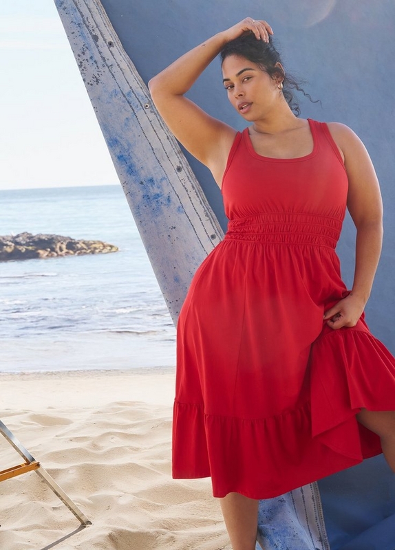 'A plus-size woman with dark hair pulled up loosely poses leaning against a weathered blue painted surfboard on a sandy beach with the ocean and rocky shore visible in the background. She wears small gold hoop earrings and a solid red sleeveless midi dress with a square racerback neckline, a wide smocked waistband, and a tiered ruffle hem skirt. She holds the skirt lightly with one hand and has one hand raised to her hair.'
