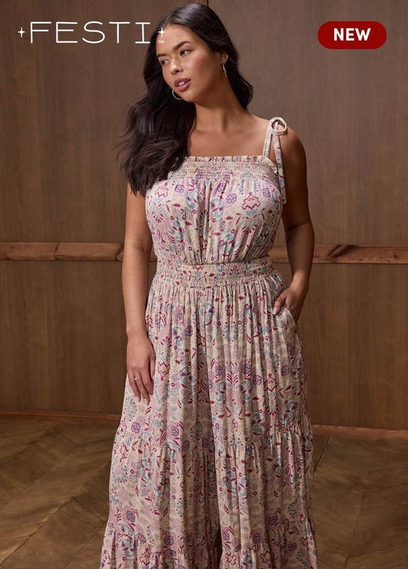 ''A plus-size woman with long dark wavy hair and gold hoop earrings poses with one hand in her pocket against a warm wood-paneled wall with herringbone wood flooring. She wears a cream and blush tiered maxi dress printed with an all-over boho floral and paisley pattern in pink, lavender, and teal, featuring a square smocked neckline, smocked waistband, wide tie straps with bow detail at the shoulders, and a flowing tiered skirt with a ruffle hem. She wears a gold ring. White text in the upper left reads '+FESTI+ TM' and a dark red rounded badge in the upper right reads 'NEW' in white text.'