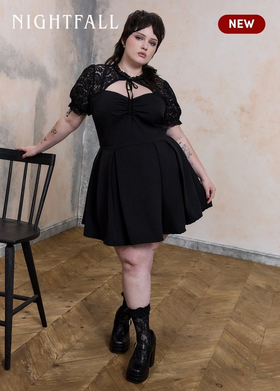 ''A plus-size woman with dark hair with fringe bangs poses standing next to a black wooden chair on herringbone wood flooring against a weathered cream and peach textured wall. She has blue eye makeup and tattoos on both forearms. She wears a black fit-and-flare mini dress with a sweetheart neckline, a gathered center-front tie detail, a cutout below the neckline, short puff sleeves in sheer black floral lace with ruffled cuffs, and a full pleated skirt. She is paired with black chunky platform lace-up ankle boots and black lace ruffle ankle socks. White serif text in the upper left reads 'NIGHTFALL' and a dark red rounded badge in the upper right reads 'NEW' in white text.'