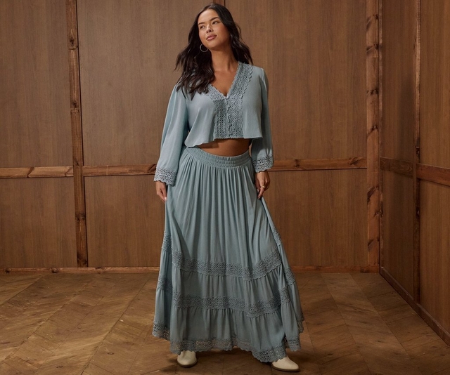 A woman with long dark wavy hair stands in a room with warm brown wood-paneled walls and a chevron parquet floor, looking off to one side. She wears gold hoop earrings and a dusty teal two-piece set consisting of a cropped long-sleeve top with a V-neckline, button front, and crochet lace trim at the neckline, hem, and cuffs, paired with a flowing tiered maxi skirt in the same color with horizontal crochet lace insets and a smocked waistband. She wears white ankle boots and a ring on her right hand.