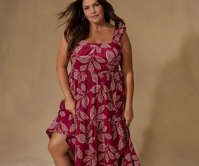 A woman with long dark wavy hair and dangling earrings stands against a soft warm beige backdrop, looking directly at the camera. She wears a sleeveless tiered dress in deep magenta with an allover cream botanical leaf print. The dress has a square neckline with gathered fabric, a ruffle strap on one shoulder, a smocked empire waist, and tiered skirt panels. Both hands rest near the pockets, slightly gathering the skirt. The image is cropped above the knee.