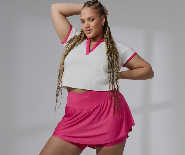 'A plus-size woman with long highlighted box braids poses with one arm raised behind her head against a light gray background. She wears small gold hoop earrings and a white cropped short-sleeve polo-style top with hot pink contrast trim at the collar, sleeves, and seams, paired with a hot pink athletic skort featuring a smooth high waistband and a ruffled skirt overlay with built-in shorts underneath.'