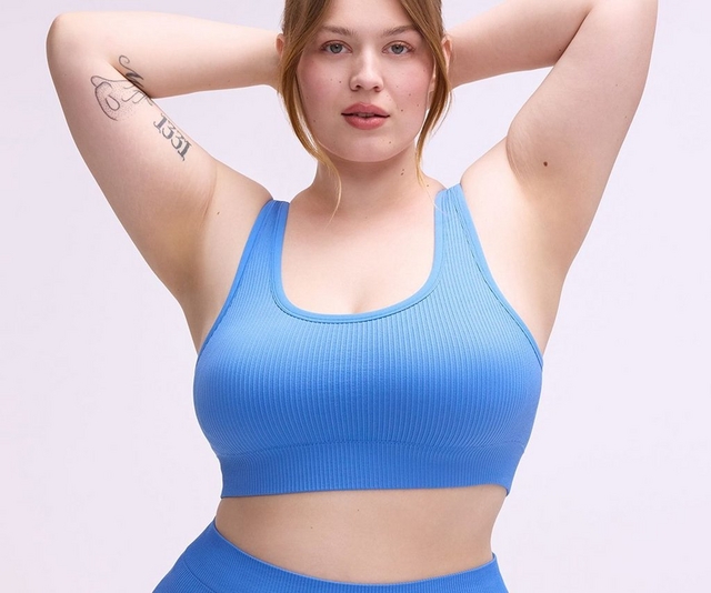 'A plus-size woman with strawberry blonde hair pulled back loosely poses with both arms raised and hands clasped behind her head against a soft lavender-gray background. She has tattoos on her upper inner arm and lower leg, and wears a matching periwinkle blue ribbed seamless lingerie set consisting of a scoop-neck racerback sports bra crop top and high-waist full-coverage briefs with a smooth waistband.'