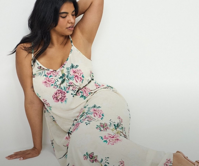 'A plus-size woman with long dark hair sits on the floor leaning against a bright white wall, one arm raised with her hand resting behind her head, looking downward. She wears a cream satin slip-style maxi dress with thin adjustable spaghetti straps and a V-neckline, printed with large pink peony and rose florals with green leaves and delicate botanical detailing throughout.'