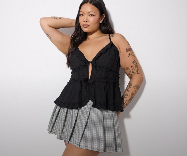 'A plus-size woman with long dark hair and extensive tattoos on both arms poses with one arm raised behind her head against a white wall. She wears small gold hoop earrings, a black textured spaghetti strap camisole top with a deep V-neckline, a keyhole cutout at the center front with a small tie detail, and multiple rows of ruffle trim detailing in a tiered peplum silhouette, paired with a black and white micro houndstooth pleated mini skirt.'