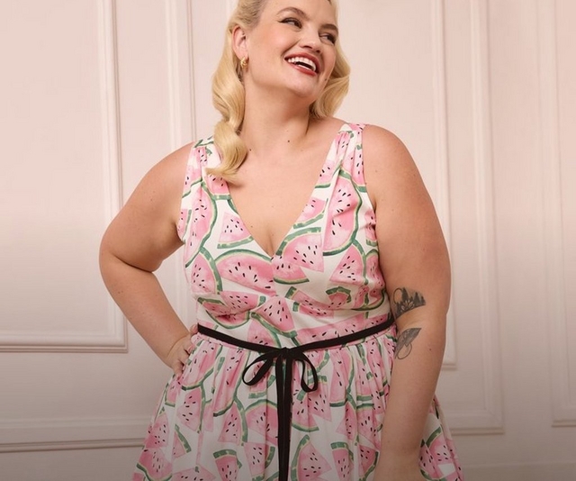 'A plus-size woman with short wavy blonde hair and red lipstick laughs and looks to the side while standing against a soft blush pink paneled wall. She wears small gold earrings and a sleeveless white fit-and-flare midi dress with a deep V-neckline, printed with large pink watermelon slice illustrations with green rinds and black seeds, cinched at the waist with a black velvet ribbon tie bow. She has a tattoo visible on her right forearm and stands with both hands on her hips.'