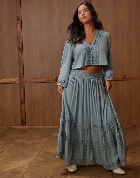A woman with long dark wavy hair stands against a warm brown wood-paneled wall, looking off to the side. She wears gold hoop earrings and a dusty teal two-piece set consisting of a cropped long-sleeve top with a V-neckline, button front, and crochet lace trim at the neckline, hem, and cuffs, paired with a flowing tiered maxi skirt in the same color featuring horizontal crochet lace insets and a smocked waistband. The skirt brushes the floor, revealing white ankle boots. She wears a ring on her right hand. The floor is a chevron-patterned light wood parquet.