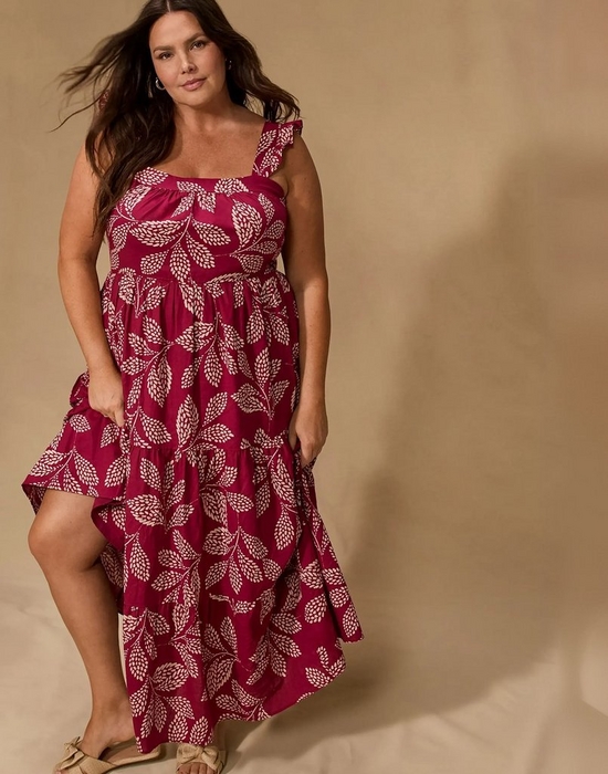 A woman with long dark wavy hair and hoop earrings stands in front of a soft warm beige backdrop, looking directly at the camera. She wears a sleeveless tiered midi dress in deep magenta with an allover cream botanical leaf print. The dress features a square neckline with gathered fabric, a ruffle strap on one shoulder, a smocked empire waist, and tiered skirt panels that fall to an asymmetrical high-low hem. Her hands rest at her sides near the pockets, and she wears tan strappy sandals with bow detail.