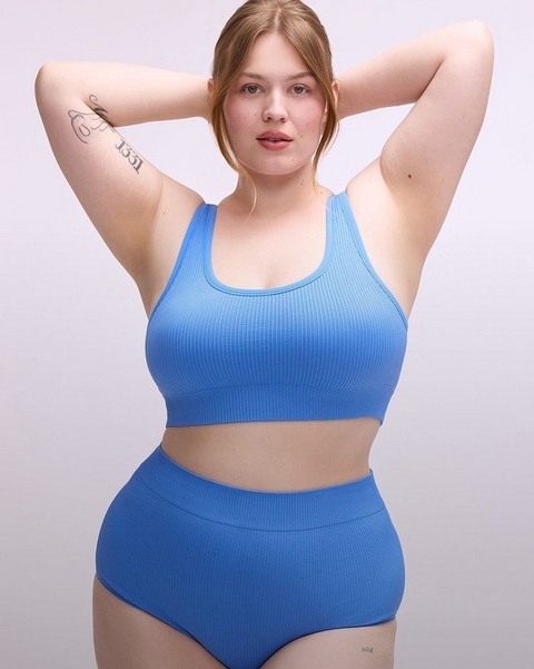 'A plus-size woman with strawberry blonde hair pulled back loosely poses with both arms raised and hands clasped behind her head against a soft lavender-gray background. She has tattoos on her upper inner arm and lower leg, and wears a matching periwinkle blue ribbed seamless lingerie set consisting of a scoop-neck racerback sports bra crop top and high-waist full-coverage briefs with a smooth waistband.''