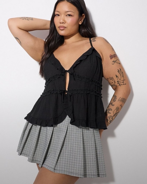 'A plus-size woman with long dark hair and extensive tattoos on both arms poses with one arm raised behind her head against a white wall. She wears small gold hoop earrings, a black textured spaghetti strap camisole top with a deep V-neckline, a keyhole cutout at the center front with a small tie detail, and multiple rows of ruffle trim detailing in a tiered peplum silhouette, paired with a black and white micro houndstooth pleated mini skirt.'