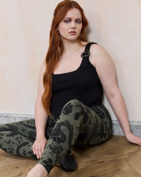 'A plus-size woman with long straight auburn hair and smoky eye makeup sits on a herringbone wood floor leaning against a weathered cream and peach textured wall with white painted baseboards. She wears small silver chain drop earrings and a silver ring, a fitted black square-neck tank top with a decorative black hardware and chain detail on one wide strap, and olive green leggings with an all-over black botanical and peacock jacquard print. She wears black flat shoes.''