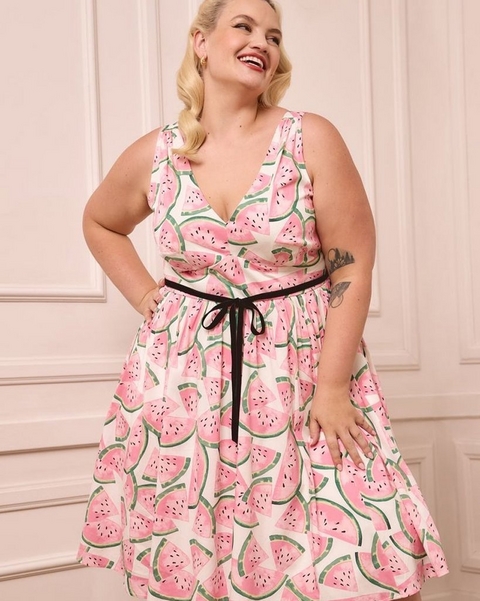 'A plus-size woman with short wavy blonde hair and red lipstick laughs and looks to the side while standing against a soft blush pink paneled wall. She wears small gold earrings and a sleeveless white fit-and-flare midi dress with a deep V-neckline, printed with large pink watermelon slice illustrations with green rinds and black seeds, cinched at the waist with a black velvet ribbon tie bow. She has a tattoo visible on her right forearm and stands with both hands on her hips.'