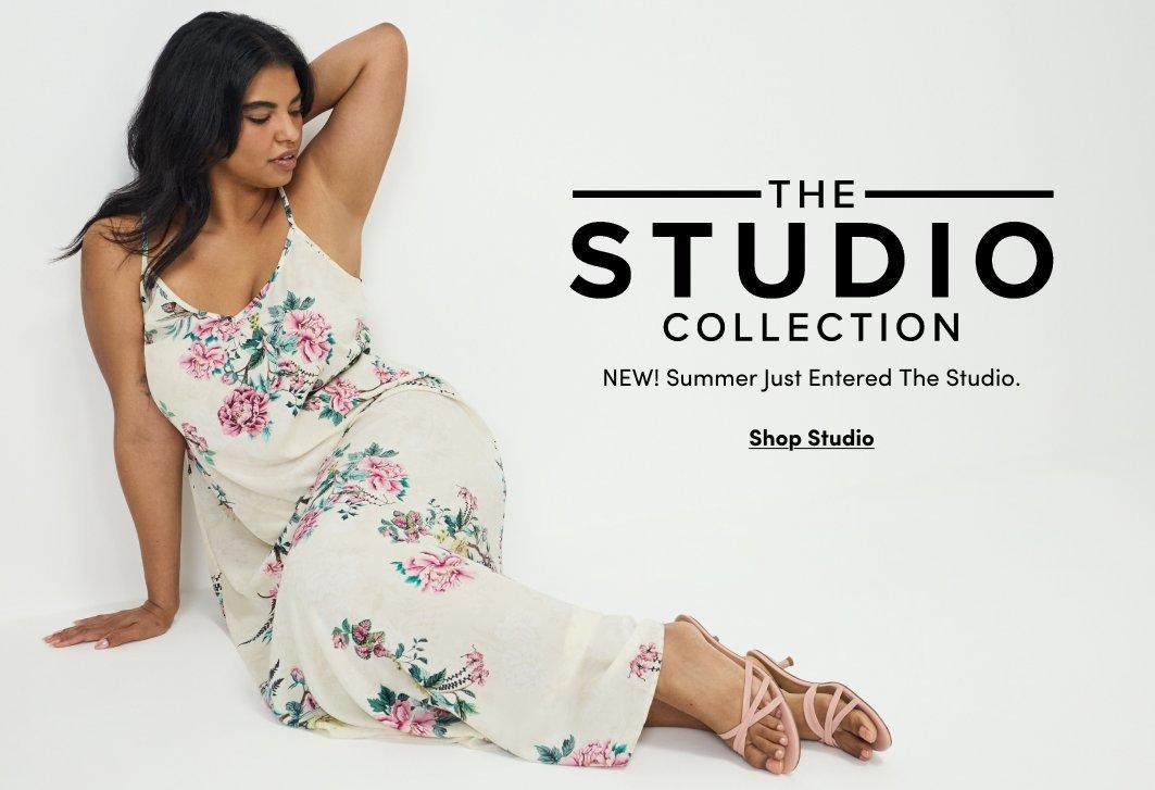 A promotional banner with a white background. On the left, a woman with dark hair pulled up sits on the floor with her legs extended, leaning on one arm. She wears a sleeveless floral maxi dress in cream with pink, green, and coral flower print, and strappy heeled sandals in a neutral tan. On the right side, black text reads 'THE STUDIO COLLECTION' with 'STUDIO' in large bold letters, followed by smaller text reading 'NEW! Summer Just Entered The Studio.' Below that is a dark rectangular button with white text reading 'Shop Studio'.